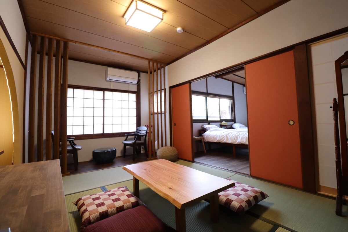 A traditional Japanese room features tatami mats and shoji screens. A low wooden table is surrounded by floor cushions. On one side, two chairs are positioned near a window, while a bed is neatly arranged in the background, enhancing the serene atmosphere.