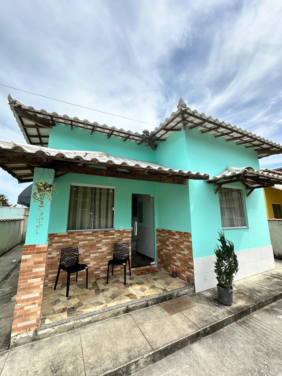 The exterior of a charming house showcases a turquoise color with a stone accent at the base. A covered porch is visible, featuring two chairs and a potted plant, creating a welcoming entryway. The roof features traditional tile detailing, enhancing the overall character of the home.