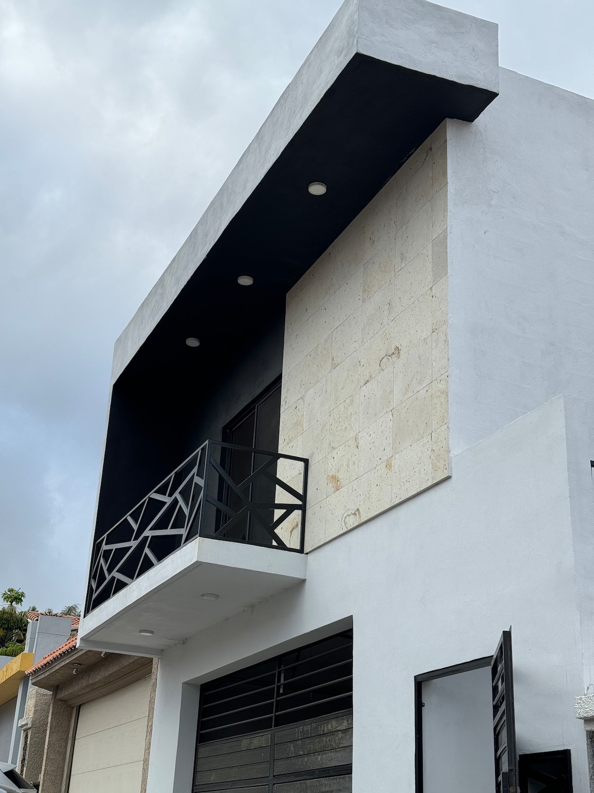 A modern building exterior is shown, featuring a sleek balcony with a geometric railing. The upper level's wall is adorned with light-colored stone, contrasting with a dark overhang. The entrance is framed by large glass doors, providing a contemporary aesthetic.