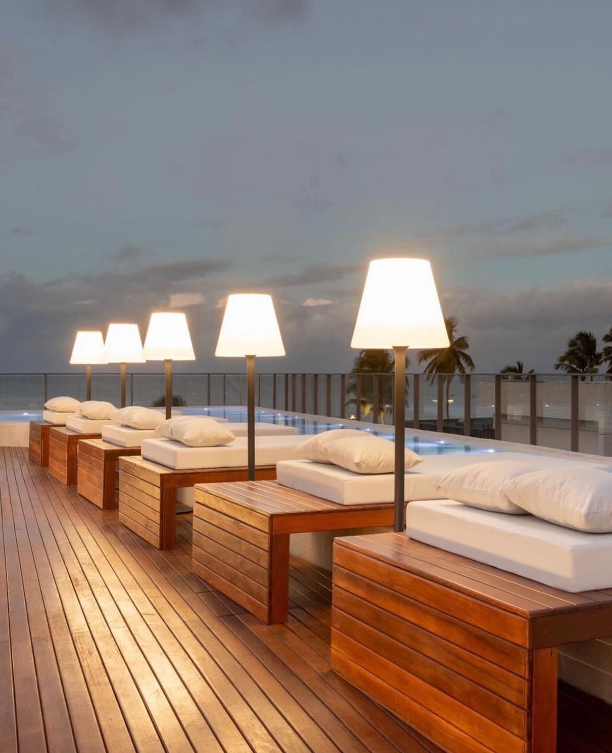 The rooftop area features several wooden lounge chairs with plush white cushions, complemented by elegant lamps that provide soft lighting. The warm-toned wooden deck enhances the inviting atmosphere, while the expansive view offers glimpses of the surrounding palm trees and ocean.