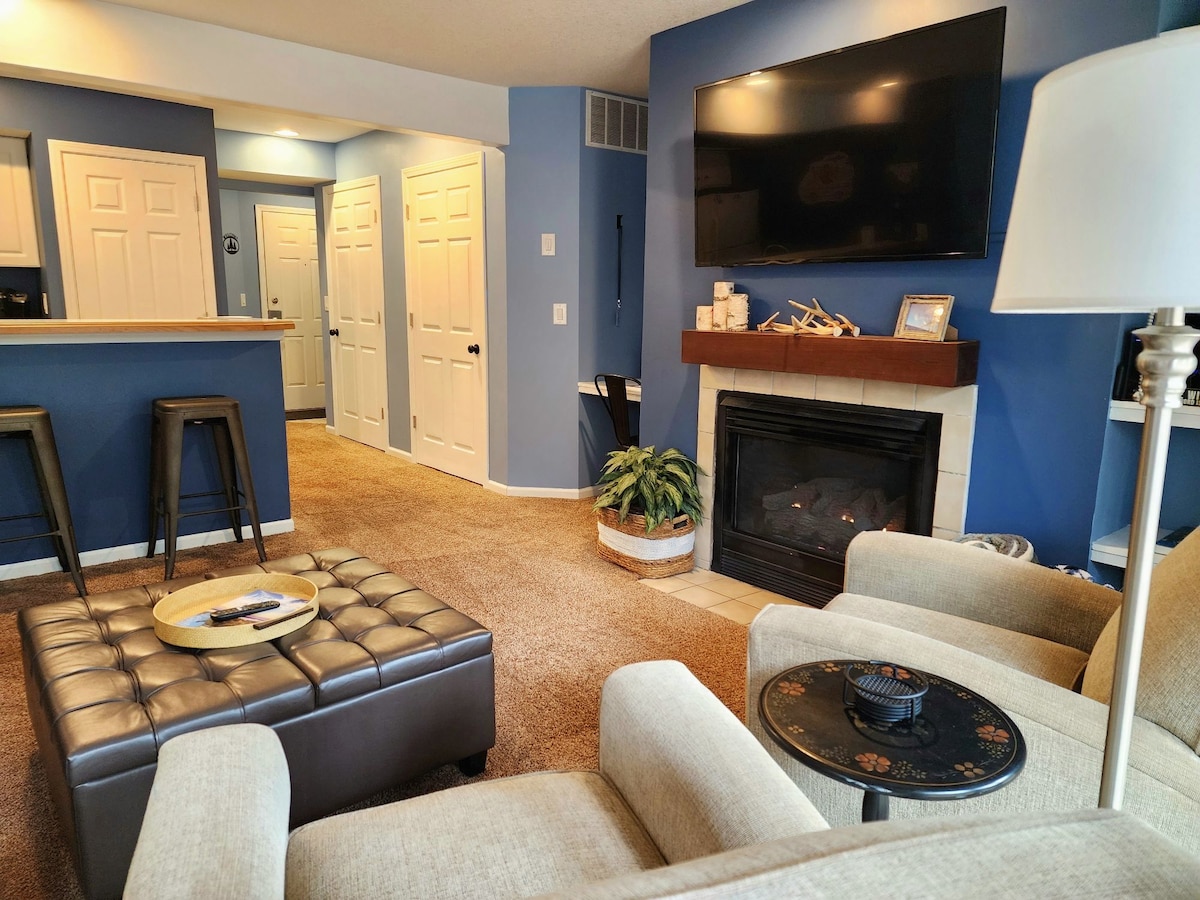 The living area features a cozy seating space with light-colored armchairs and a tufted coffee table. A wall-mounted television is above a gas fireplace. The room is painted in soft blue tones, with a view into the dining area and hallway leading to bedrooms.