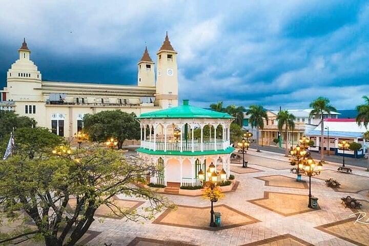 A charming plaza is showcased, featuring a round gazebo with a green roof, surrounded by trees and decorative street lamps. The backdrop includes a historical building with towers and a clock, under a cloudy sky, contributing to the tranquil atmosphere of the area.