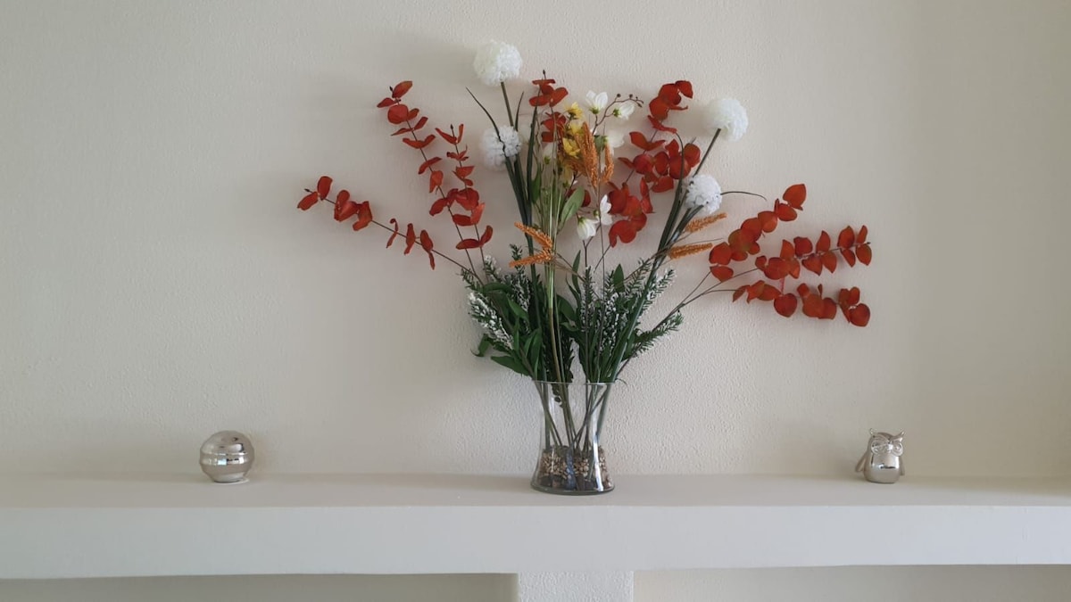 A decorative shelf features a clear vase filled with a variety of vibrant flowers in shades of red and white, complemented by greenery. Two small silver decorative items are positioned on either side of the vase against a neutral, light-colored wall.