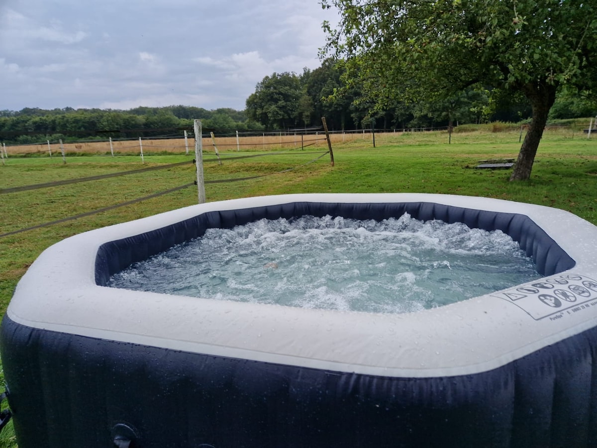 An inflatable hot tub is situated on a grassy area surrounded by a natural landscape. Gentle waves are visible on the water's surface, indicating the tub is filled and in use. Fences and trees frame the background, enhancing the serene outdoor setting.