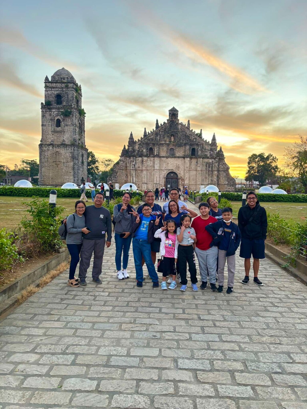 A group of individuals stands on a cobblestone pathway, framed by a historic stone church and a picturesque sunset. The church features intricate architecture, while the grassy landscape surrounds the structure, providing a natural backdrop to the scene.