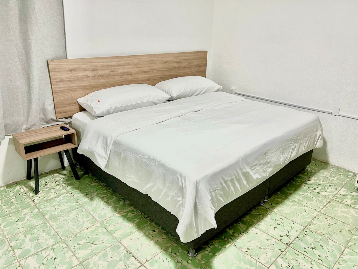 A spacious bed is situated against a light-colored wall, featuring a simple wooden headboard and two soft pillows. A small nightstand with a remote control is placed beside the bed. The floor is covered with a unique green tile pattern.