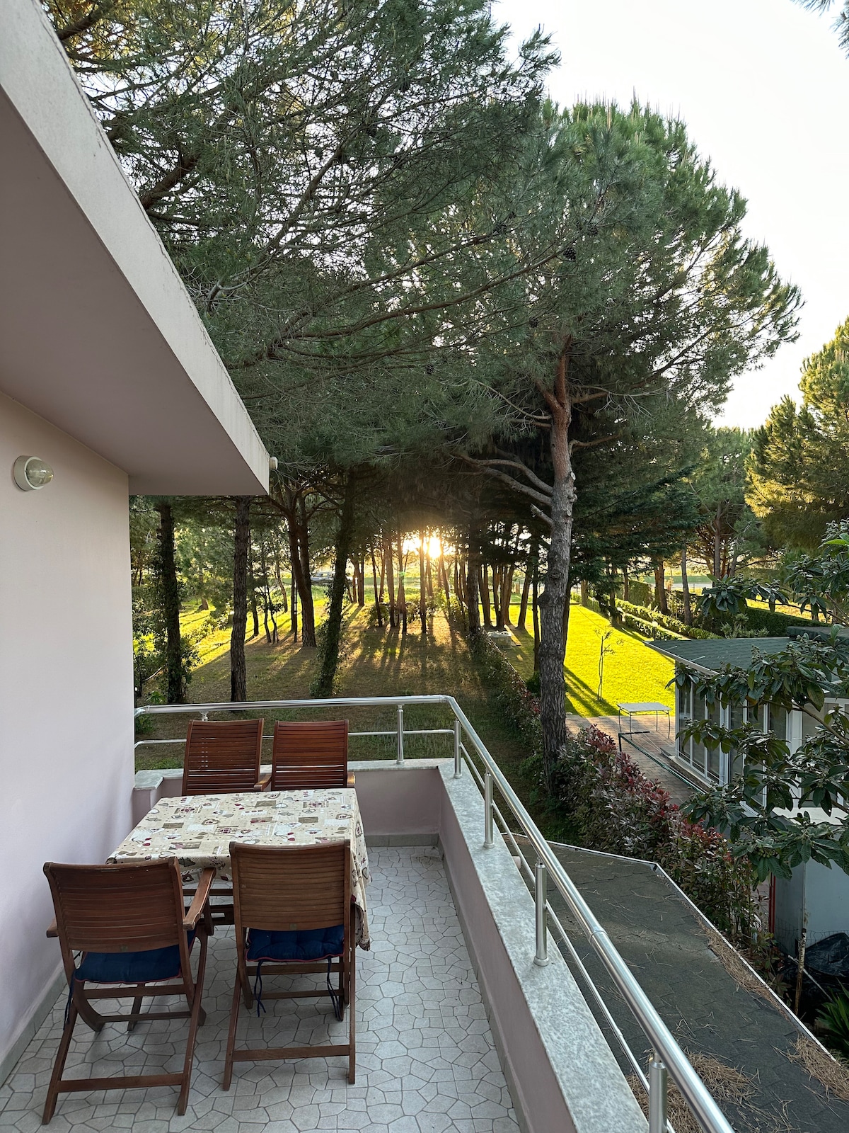 A balcony is shown with a table covered in a light cloth and four wooden chairs. Tall trees create a natural backdrop, with sunlight filtering through the branches, casting gentle shadows across the ground.