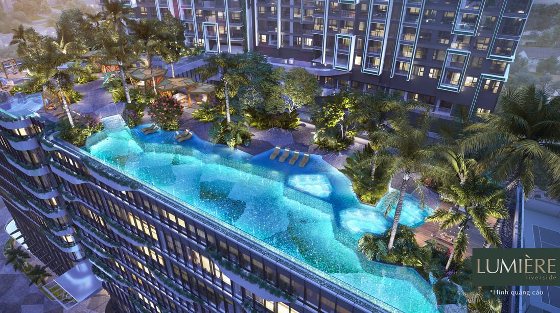 The image displays a aerial view of the Lumiere Riverside complex, showcasing an inviting outdoor swimming pool surrounded by lush greenery and palm trees. Comfortable lounge chairs are positioned along the pool edge, offering a serene atmosphere for relaxation.