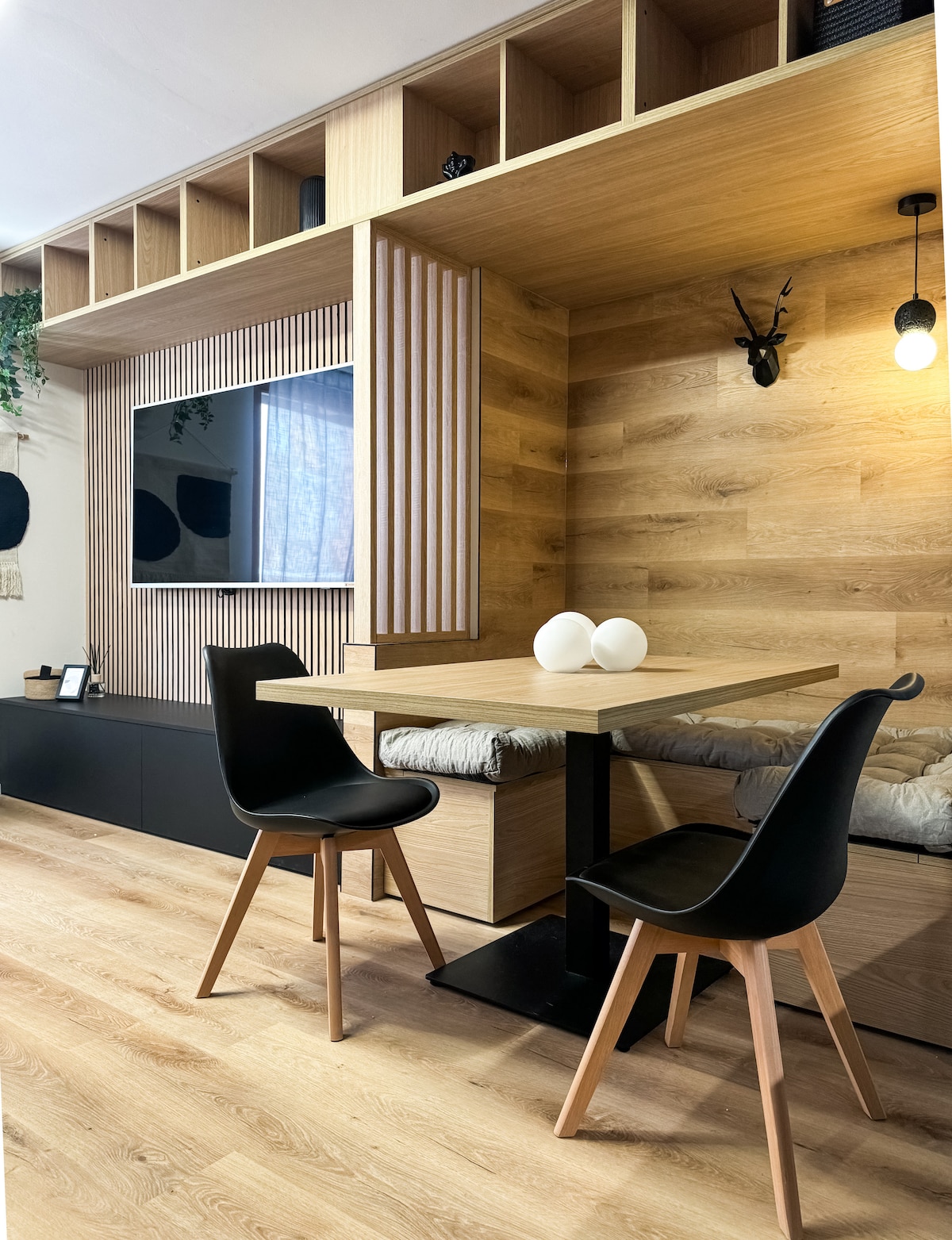 A modern living space features a wooden accent wall with built-in shelving and a mounted TV. A dining table for two is positioned in front, accompanied by two black chairs with wooden legs. Soft lighting enhances the warm atmosphere, creating a cozy setting.