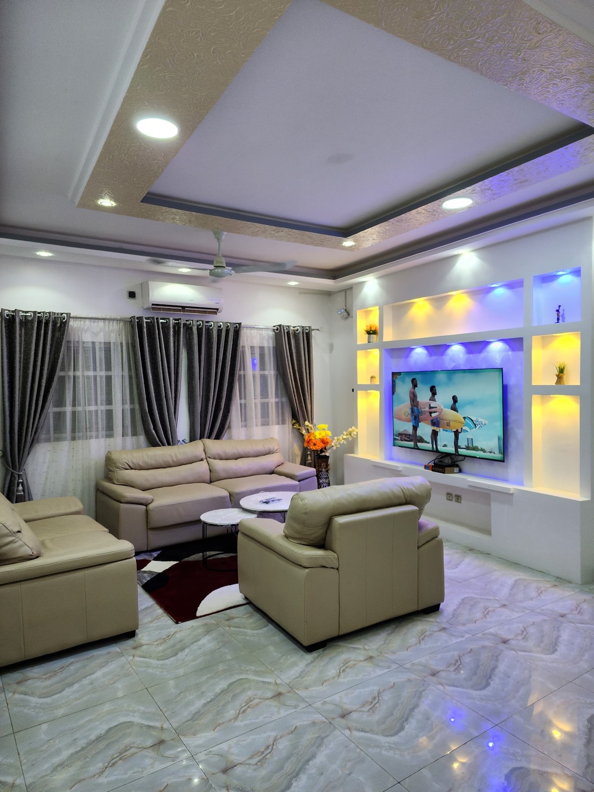 A well-lit living area features a cozy seating arrangement with light-colored sofas surrounding a circular coffee table. A large television is mounted on a wall with ambient lighting, complemented by decorative shelves. Floor-to-ceiling curtains frame the windows, enhancing the space's brightness.