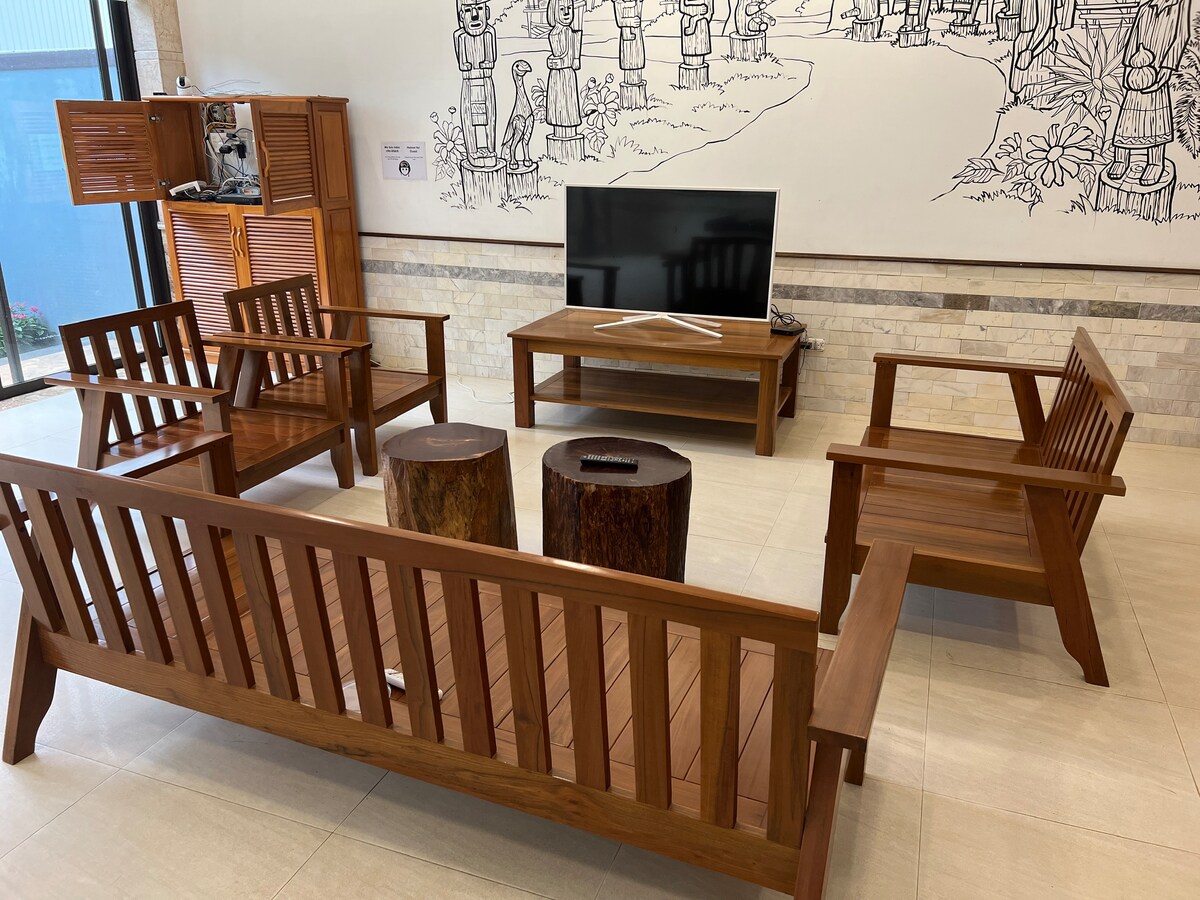 A seating area is presented with wooden benches and stools made from natural materials. A flat-screen TV is positioned on a wooden stand, set against a wall adorned with artistic drawings. The space is well-lit, featuring a neutral tile floor.