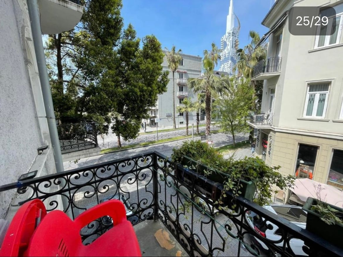 A balcony view showcases greenery from nearby palm trees and vibrant plants in pots along the railing. A red chair is visible, offering a spot to relax. The surroundings include a pedestrian-friendly street lined with buildings and a glimpse of a tall structure in the background.