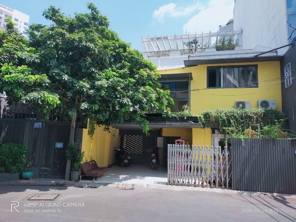 A two-story yellow building is surrounded by lush green foliage. The entrance is marked by a gated area with visible parked motorcycles. Large windows enhance the front façade, allowing ample natural light into the interior spaces.