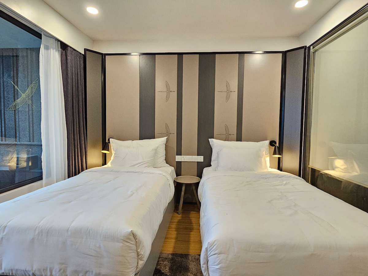 A serene sleeping area features two spacious beds dressed in crisp white linens. Soft lighting is provided by wall-mounted lamps on either side, while large windows allow natural light to fill the room, creating a bright and airy environment.