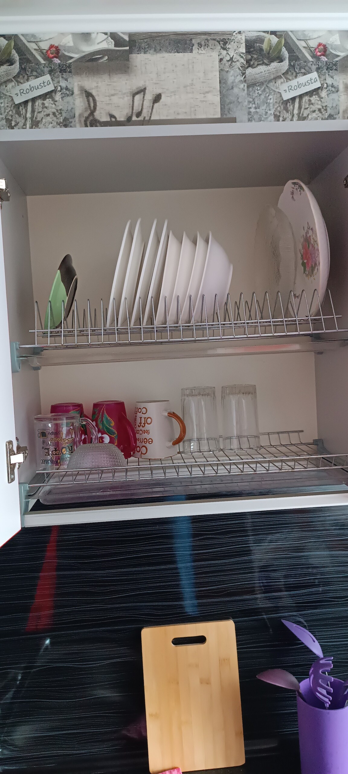 A kitchen cabinet holds organized tableware. The upper shelf features a neatly arranged stack of plates, while the lower shelf contains glasses and colorful cups. Items are stored on a drying rack, with a wooden cutting board placed on the cabinet surface below.