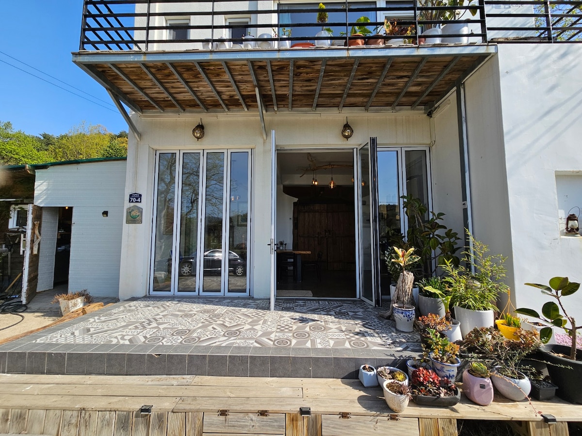 The entrance features large glass doors that open onto a tiled patio adorned with various potted plants. A wooden deck extends from the entryway, providing a connection to the outdoors. Natural light fills the space, enhancing the inviting atmosphere of the interior.