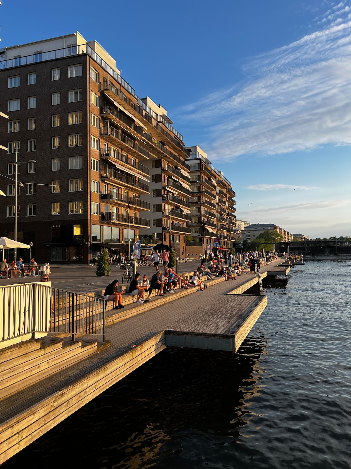 A wooden promenade lines the waterfront, where individuals enjoy the evening sun. Several multi-story buildings are visible, featuring balconies adorned with plants. The shimmering water reflects the golden hues of the sunset, creating a serene atmosphere along this vibrant urban space.