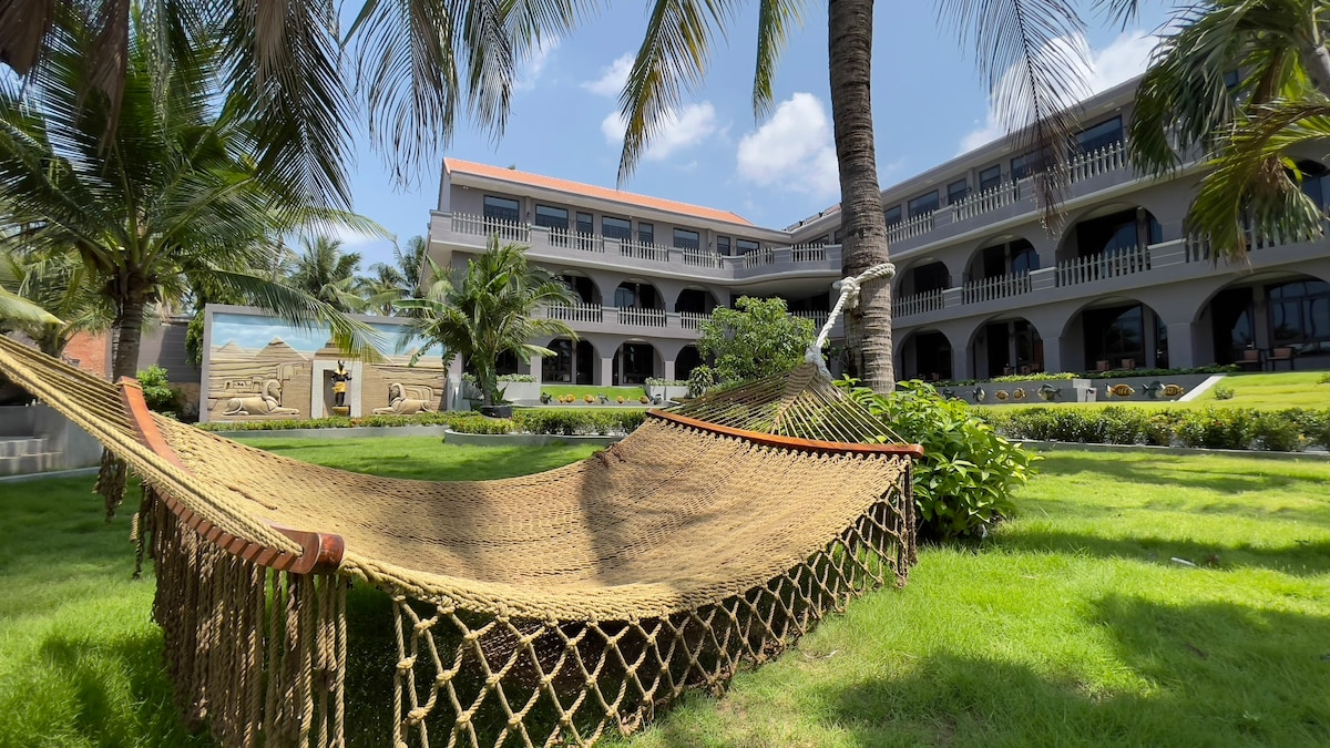 A hammocks are positioned under palm trees, set within a spacious green lawn. The resort building, featuring multiple arches and balconies, is visible in the background, surrounded by tropical landscaping.