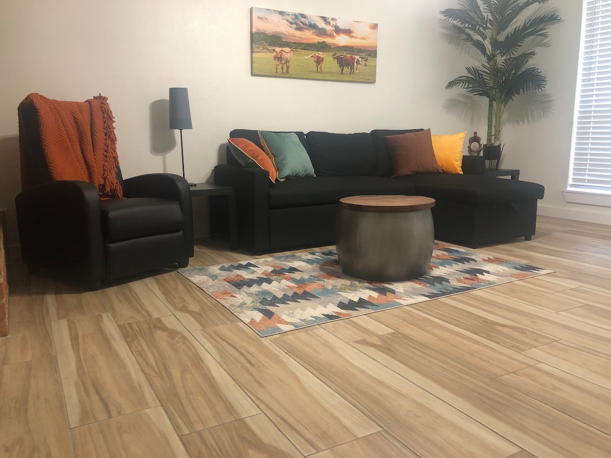 The living area features a comfortable black sofa adorned with colorful cushions, accompanied by a recliner chair. A round wooden coffee table sits on a patterned area rug over light-colored tiled flooring. A floor lamp provides illumination, and a decorative plant adds a touch of greenery.