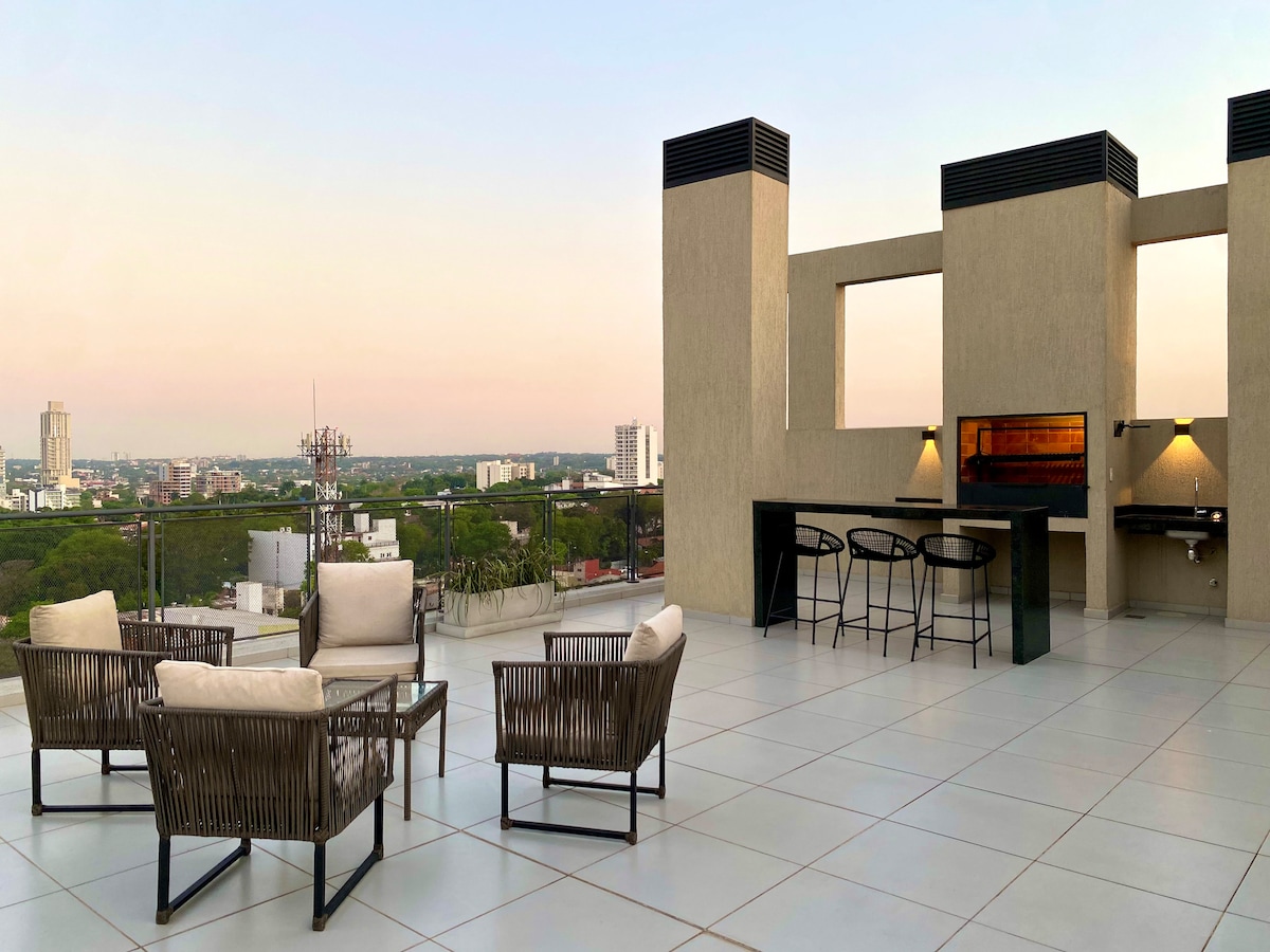 An open rooftop terrace features comfortable seating arrangements with wicker chairs and soft cushions. A dining area is equipped with a bar-style counter and sleek stools, complemented by a grill station. Expansive city views are visible through large, unobstructed openings.