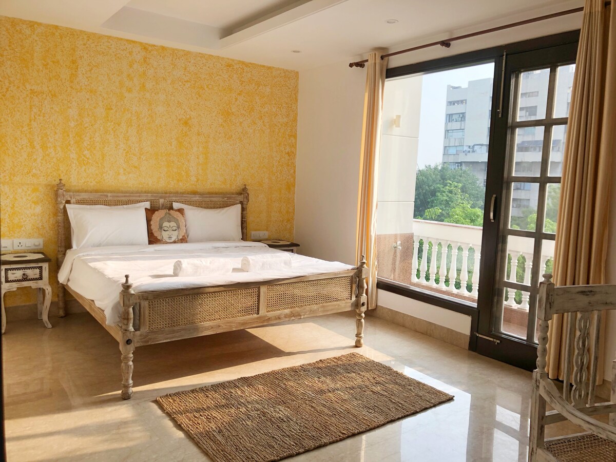 A well-lit bedroom features a spacious bed adorned with white linens. A textured yellow accent wall adds warmth to the space. Large glass doors open to a balcony, allowing natural light and a view of the greenery outside. A woven rug complements the room's decor.