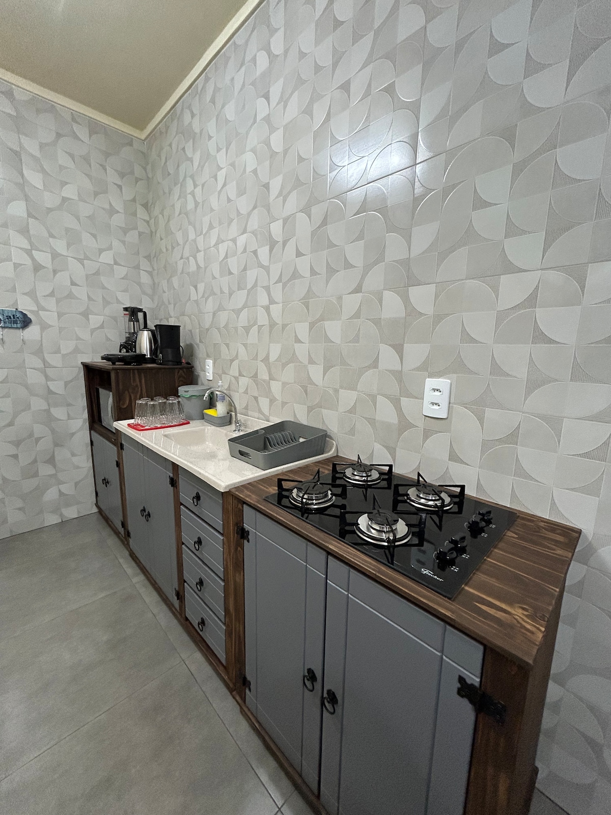 The kitchen features a modern design with patterned gray walls and a sleek countertop. A four-burner gas stove is positioned alongside storage cabinets. Kitchen essentials, including a coffee maker and a toaster, are visible on the counter, providing a functional cooking space.