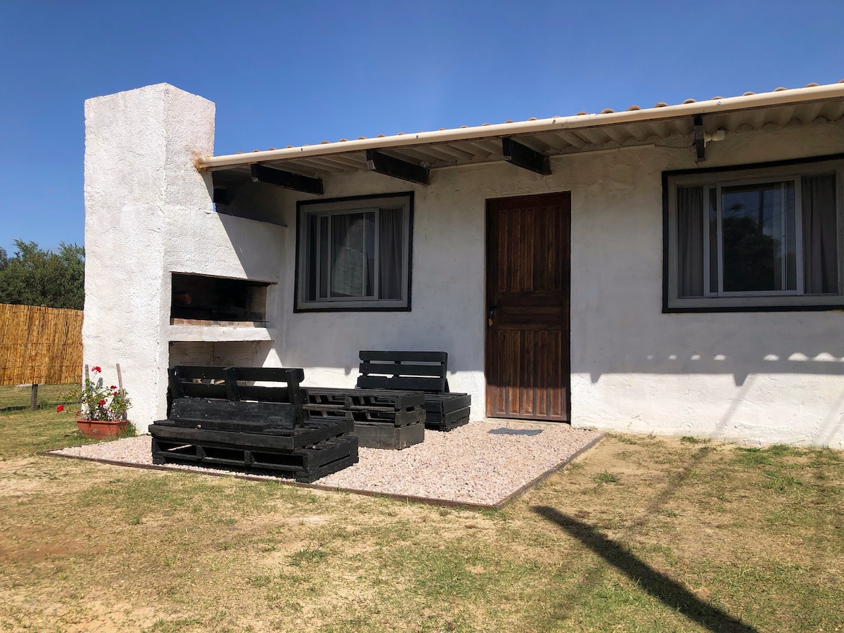 A quaint exterior view of the house features a wooden door and large windows framed with dark accents. Outdoor seating made of black pallets is arranged on a gravel surface, accompanied by a built-in grill. Lush grass surrounds the area, adding to the natural setting.
