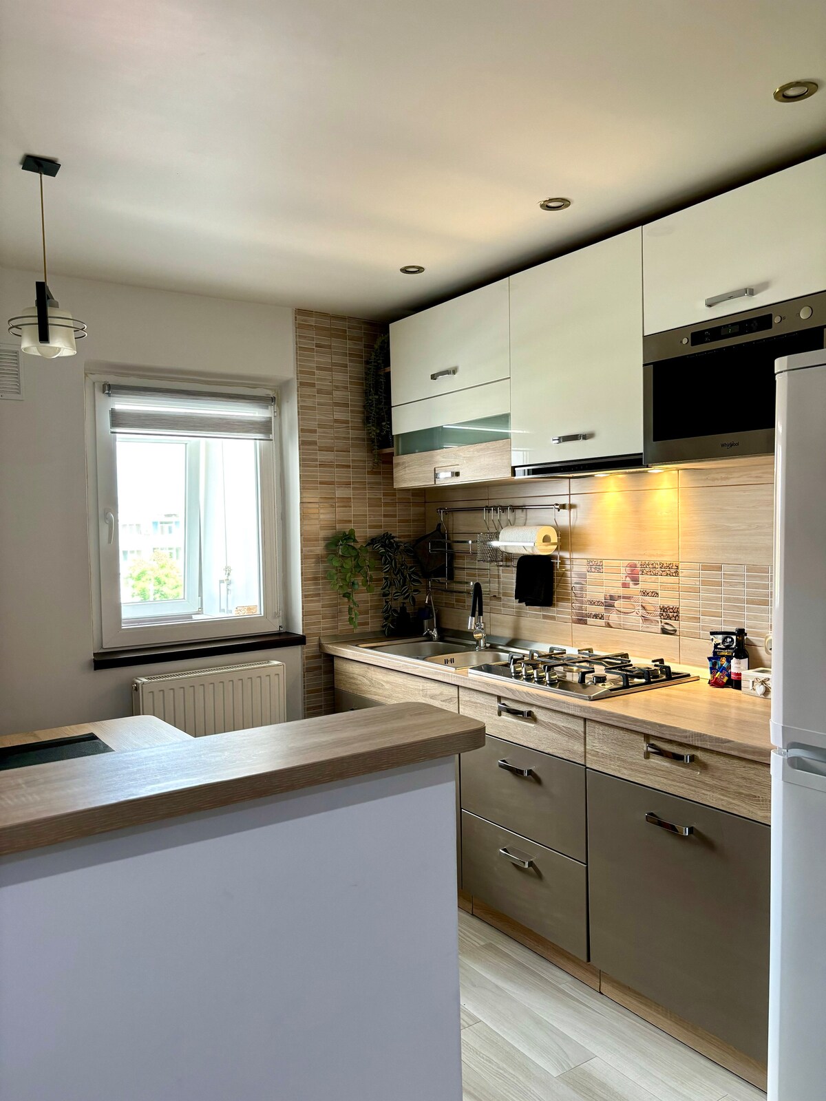 The kitchen area features modern cabinetry with light wood accents and a sleek design. A window allows natural light to brighten the space. An organized layout includes a stove, oven, and essential appliances, creating a functional cooking environment with ample countertop space.