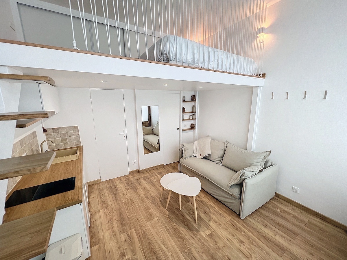 The interior features a cozy living area with a light-colored sofa, a small round table, and a kitchenette with open shelving. A mezzanine sleeping area is visible above, with a bed positioned neatly against a white wall. Natural light floods the space through large windows.