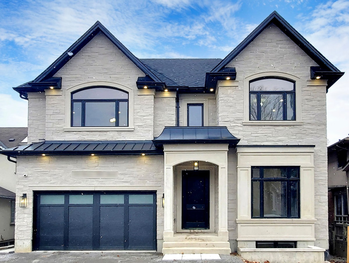 The exterior of the newly built custom home showcases a modern design with a mix of light-colored stone and dark accents. Large windows provide ample natural light, and the double garage is featured prominently beneath a stylish entrance. A clear sky is visible above.