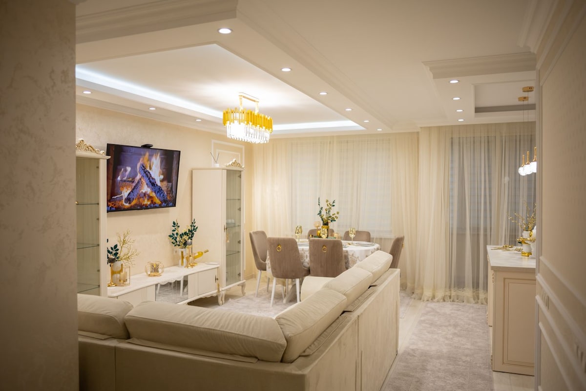 A modern living area features a cozy sectional sofa and a dining table. A flat-screen TV displays a calming fireplace scene, while soft lighting illuminates the room. Large windows with sheer curtains allow natural light to filter in, creating a serene ambiance.