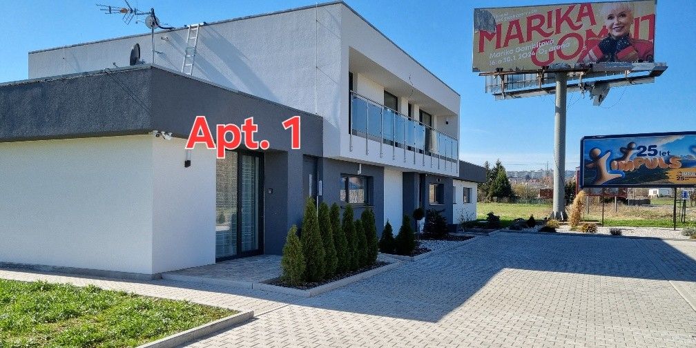 A modern two-story building features a clean facade with a balcony on the upper level. The ground level displays an entrance to Apt. 1, with well-maintained greenery and a paved parking area visible in front. A large billboard adds context to the location.