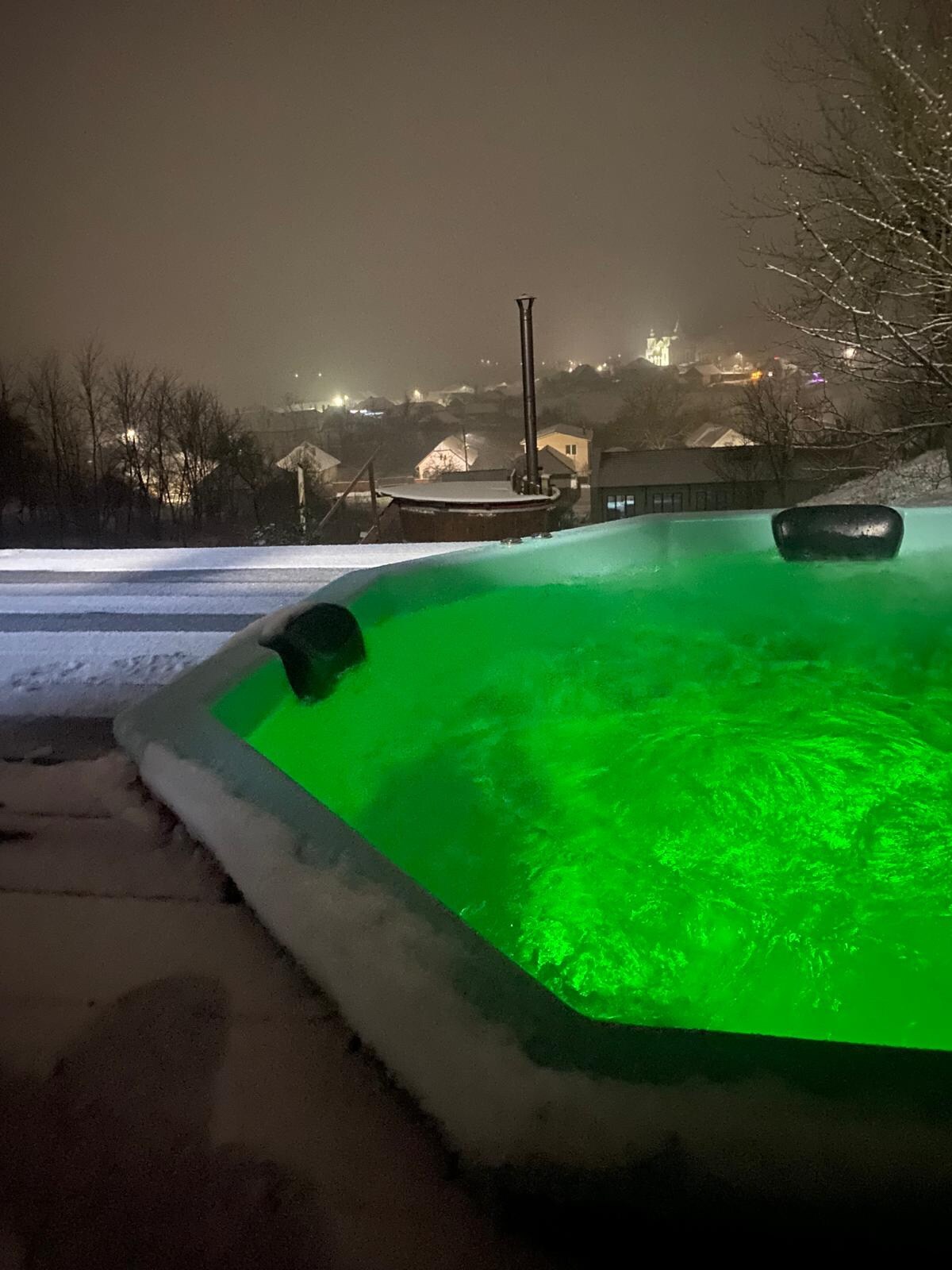 An outdoor jacuzzi is surrounded by snow, featuring soothing green lights that illuminate the water. The backdrop includes distant lights from a village, creating a serene winter scene. The setting conveys a peaceful escape, highlighted by the tranquil atmosphere.