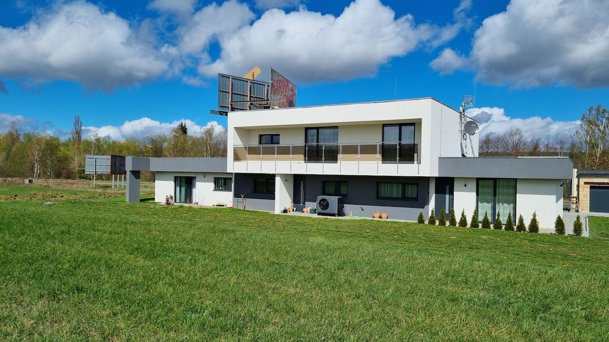 A modern two-story apartment building is set against a blue sky with scattered clouds. The well-manicured lawn surrounds the structure, which features large windows and a balcony. A grass area with small shrubs is visible in the foreground, emphasizing the spacious outdoor setting.