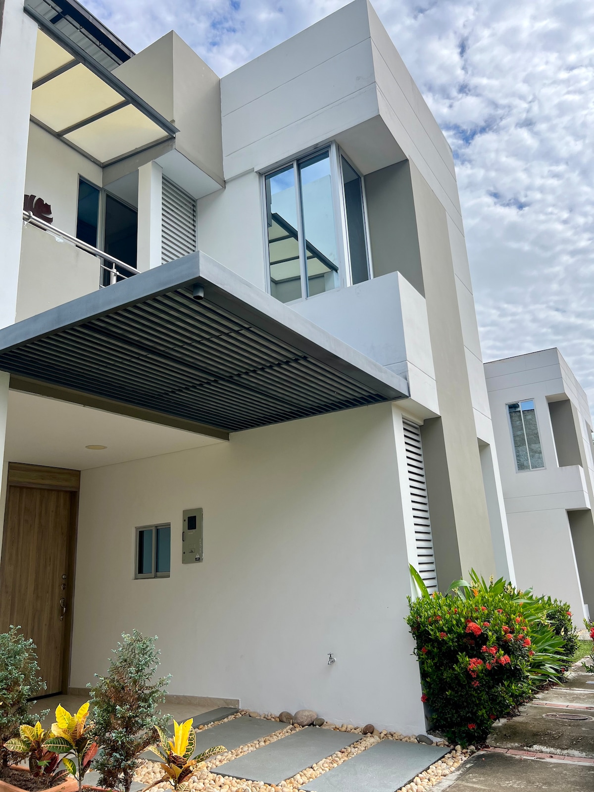 The exterior view features a modern house with a sleek design and large windows. An entrance is framed by a covered porch, while the surrounding area includes manicured landscaping and decorative plants. Natural stone pathway leads to the door, enhancing the inviting entrance.