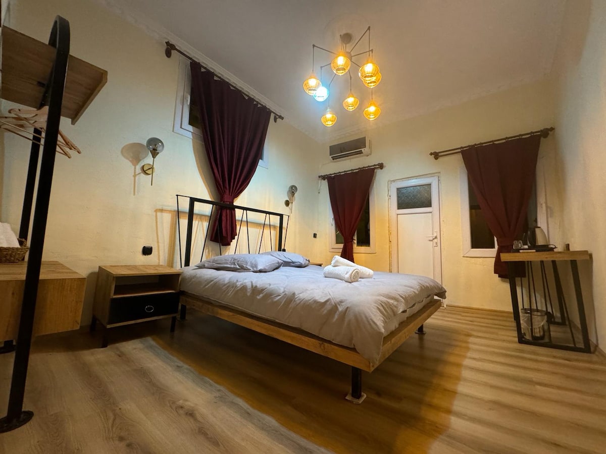 A sleeping area features a spacious bed with light bedding and a wooden frame. Two bedside lamps provide illumination, complemented by pendant lights above. Dark curtains frame the windows, and wooden flooring adds warmth to the room. A small desk is visible in the corner.