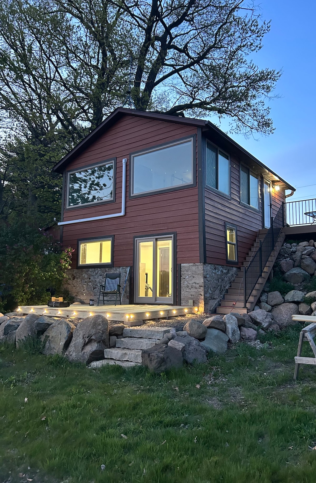 The exterior of the cabin features a modern architectural design with large windows offering ample natural light. Soft outdoor lighting illuminates the deck area, while stone accents complement the vibrant siding. A gentle slope leads up to the entrance, framed by greenery and a grassy lawn.