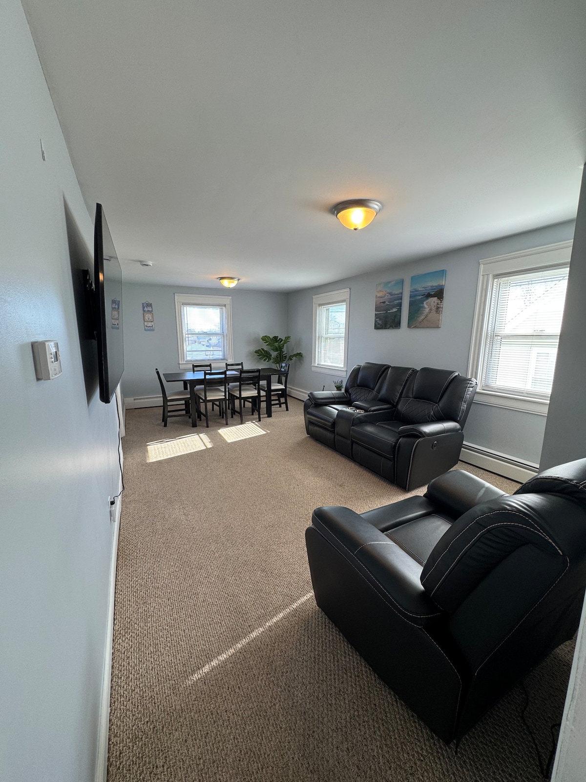 The living room features light blue walls and cozy carpeting. A black leather sofa faces a wall-mounted TV. A dining table for four is positioned near bright windows, allowing natural light. Sunlight creates patterns on the floor, enhancing the space's inviting ambiance.