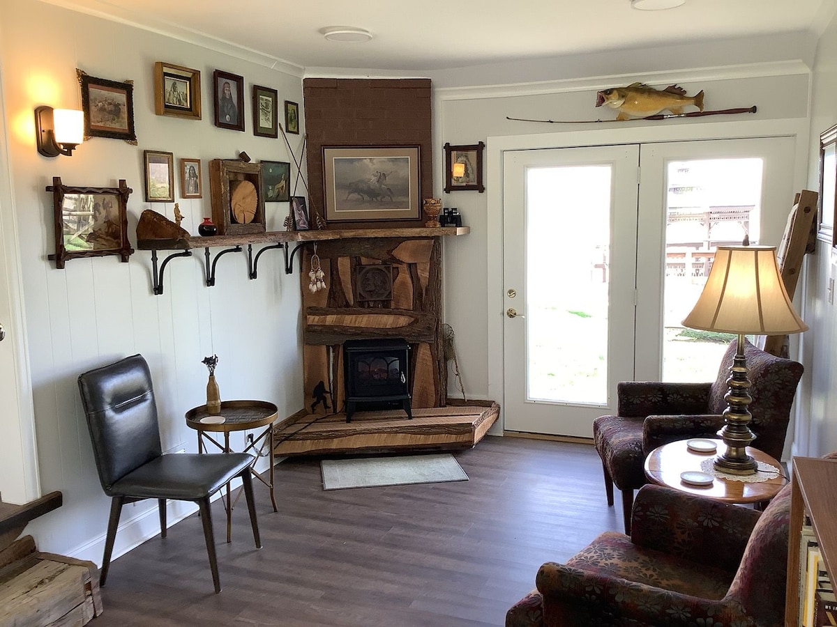 A cozy living area features a stone fireplace with a wooden mantel. Framed artwork and photographs decorate the walls. Two armchairs, one upholstered and one sleek, flank a small side table. Natural light enters through a glass door, leading to the outdoor environment.