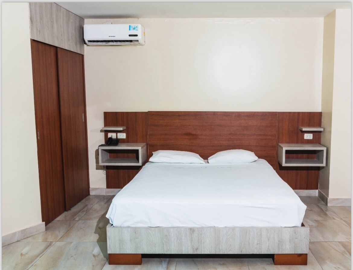 A spacious bedroom features a wooden headboard and a neatly made bed with white linens. Nightstands are positioned on either side of the bed, while soft lighting illuminates the space. A built-in closet can be seen in the background, contributing to the room's functionality.