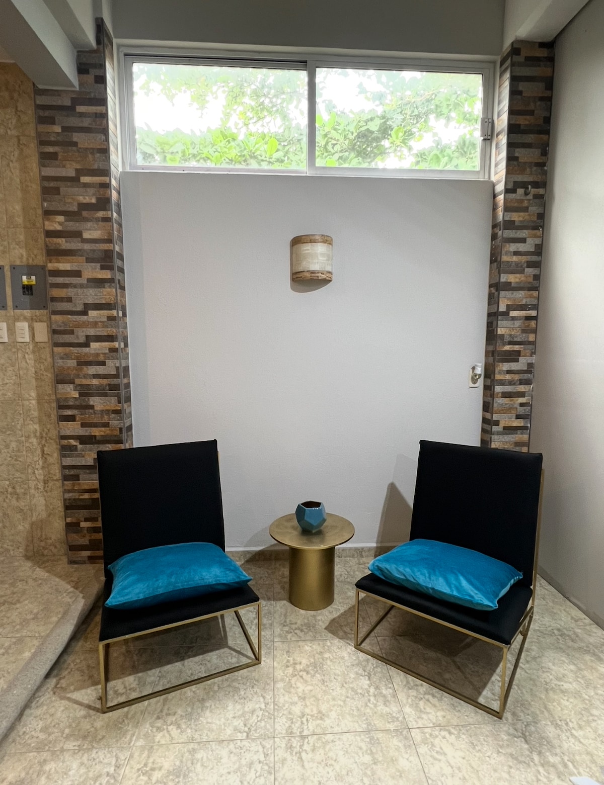A cozy corner features two black chairs with teal cushions placed around a small round table. A decorative wall light is mounted above, and a window allows natural light to illuminate the space while providing a view of greenery outside.