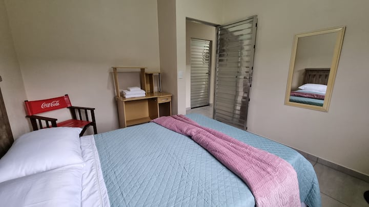 Cama de casal, cadeira de apoio, criado mudo, ventilador e pequeno armário.