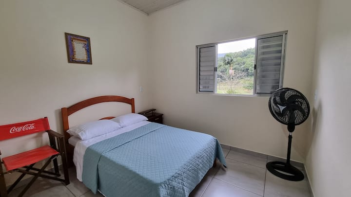 Cama de casal, cadeira de apoio, criado mudo e ventilador.