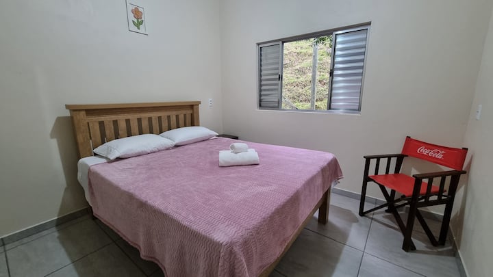 Cama de casal, cadeira de apoio, criado mudo e ventilador.