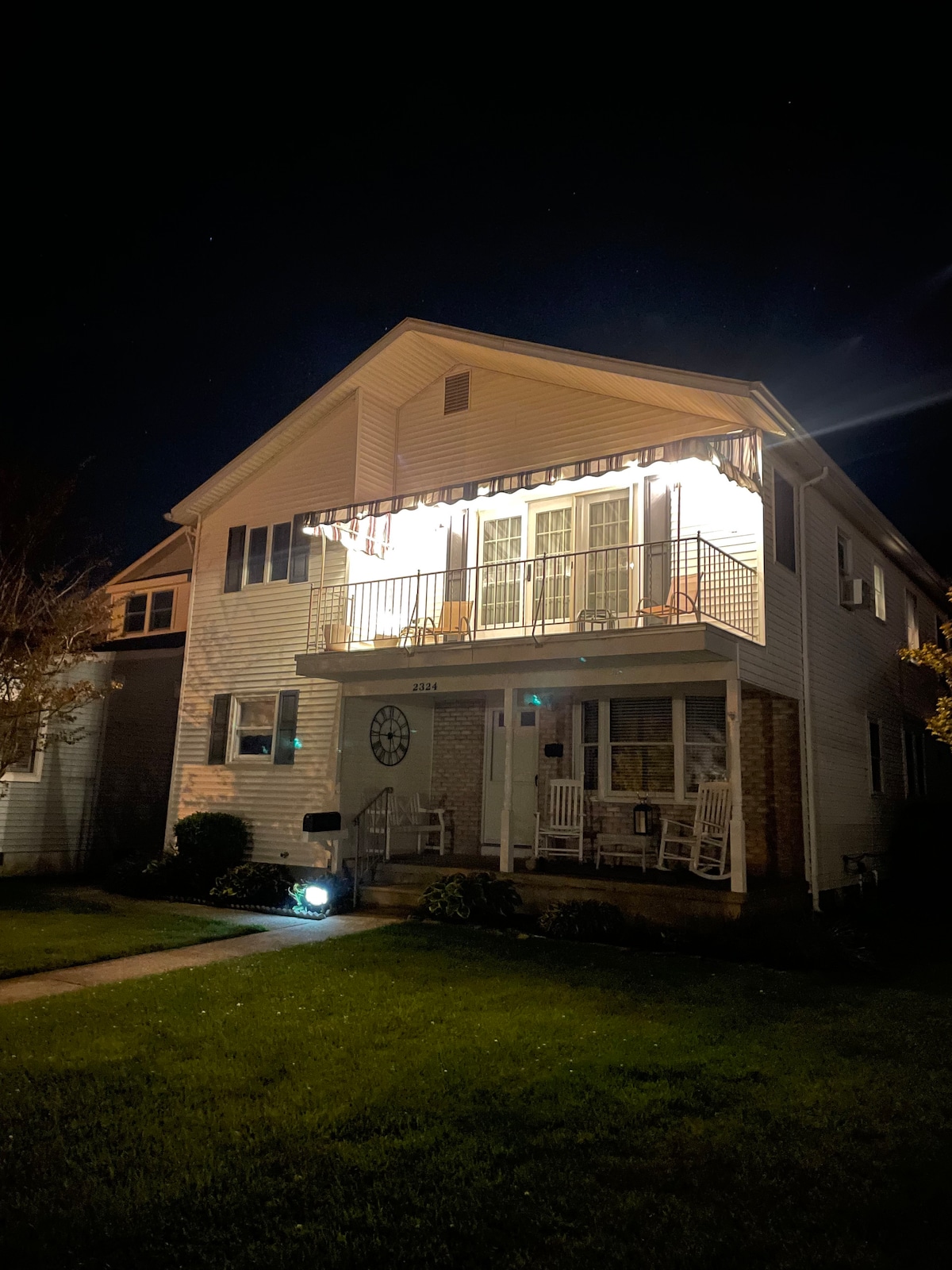 The exterior of the two-story apartment is illuminated at night, highlighting a welcoming front porch with white rocking chairs. A balcony is visible on the second floor, adorned with light-colored awnings. Lush greenery surrounds the entryway, enhancing the inviting curb appeal.
