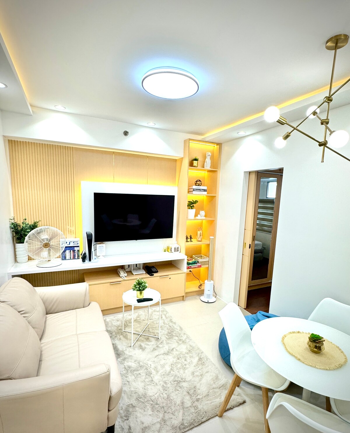 A modern living area features a light-colored sofa and a circular table with white chairs. The wall-mounted television is complemented by built-in shelves displaying decorative items. Soft light is provided by a ceiling fixture, while an electric fan is positioned nearby for ventilation.