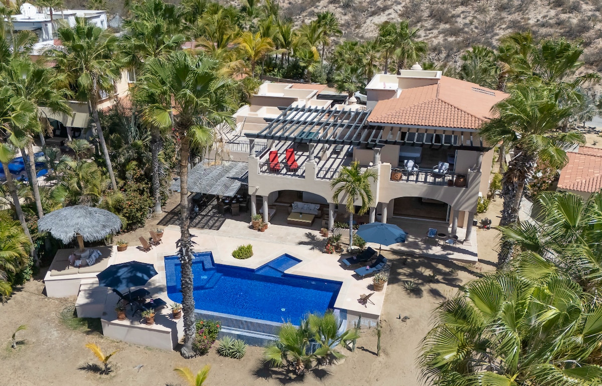 An aerial view captures the villa's expansive and landscaped surroundings. The inviting infinity pool is framed by palm trees, with a shaded lounging area nearby. The poolside terrace features outdoor seating, and the upper level showcases a spacious roof terrace ideal for relaxation.