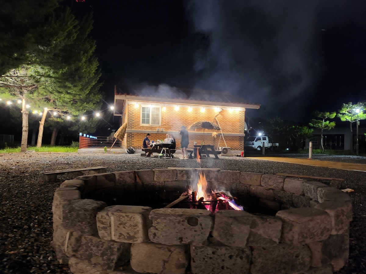 A stone fire pit is illuminated by a warm glow as flames rise, providing a focal point in the foreground. A cozy cabin is visible in the background, lit by exterior string lights, with outdoor seating and a grill situated nearby.