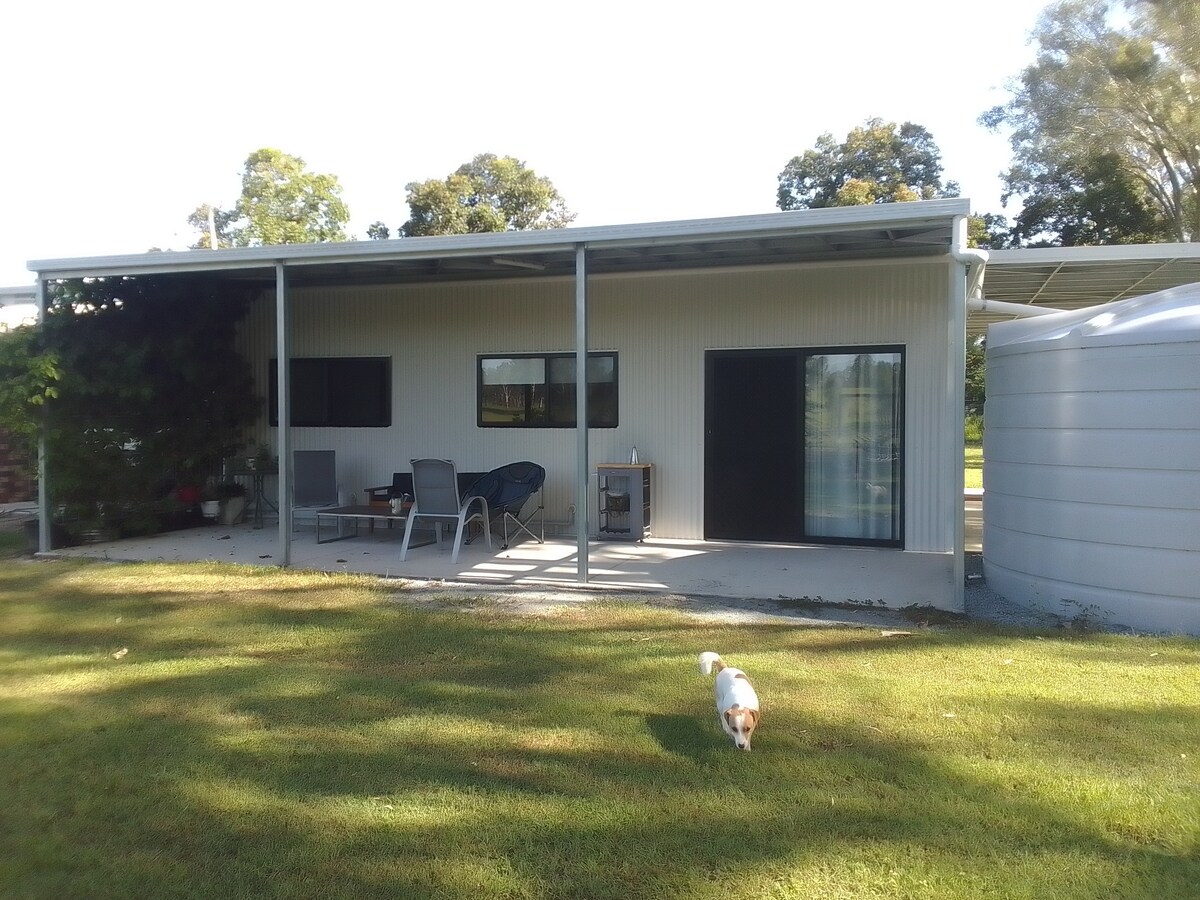 The exterior of the cabin is visible, featuring a covered patio area with seating arrangements, including chairs and a table. A small dog is seen walking on the grassy lawn. Large windows provide a view into the unit, complemented by a water tank nearby.