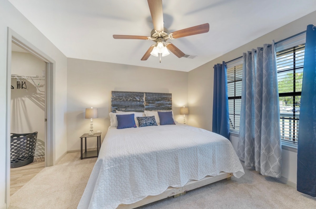 A tranquil bedroom is highlighted by a large bed with a textured headboard and decorative pillows. Two bedside lamps provide soft lighting, while a ceiling fan enhances airflow. Natural light fills the room through two windows, dressed in light curtains, and a closet is visible in the background.
